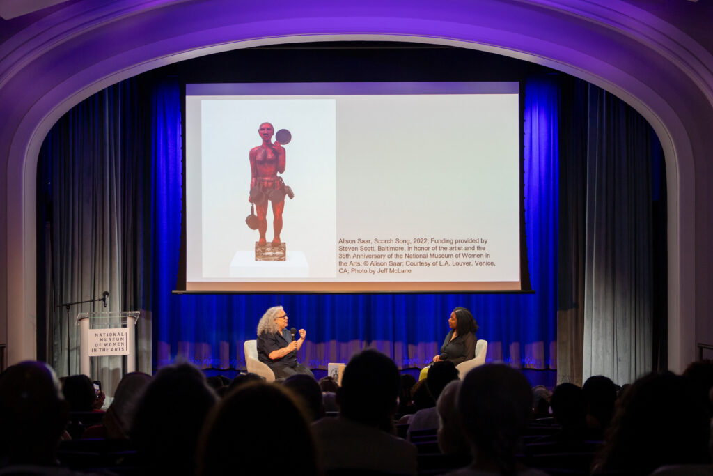 Two women sit on stage in armchairs under an arched ceiling, engaging in a discussion. A slide with an image of a red sculpture and text is projected behind them. An audience sits facing the stage.