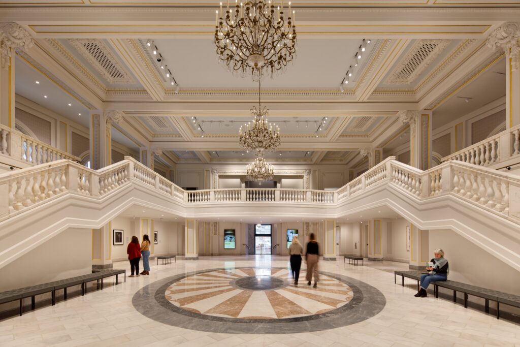A large, grand space is photographed from a wide angle. Two staircases, leading to a mezzanine level, flank a central room with an ornate marble floor. Large crystal chandeliers hang from the high ceilings.